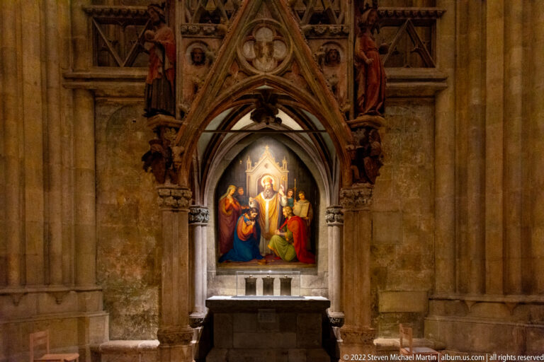 Painted religious altarpiece under a pointed Gothic stone canopy above a small altar in Regensburg Cathedral.