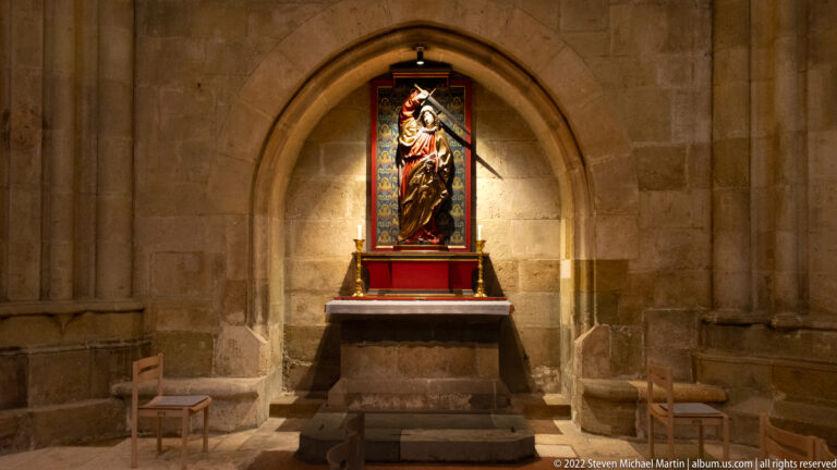 Illuminated standing saint figure in a narrow altarpiece, set in a pointed stone recess above a small altar.
