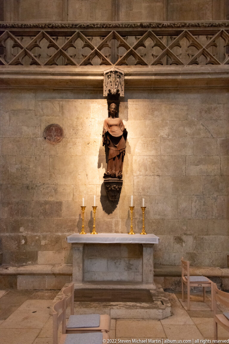 Dark crowned carved figure mounted on a stone wall above a simple altar with candlesticks and liturgical vessels.