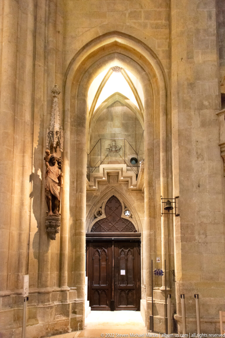 Pointed Gothic doorway with dark wooden doors, deep stone jambs, bright exterior light, and a carved figure mounted beside it.