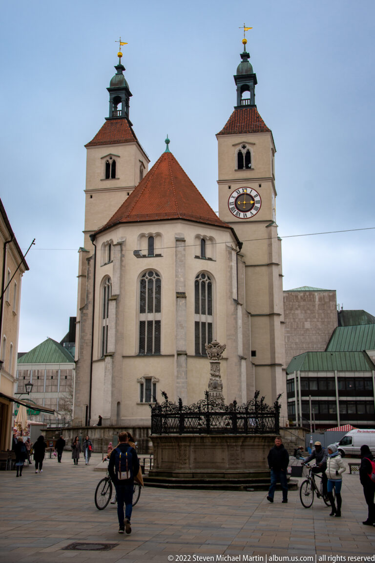 Twin-towered pale stone church facing a paved square, with clock, arched openings, large windows, pedestrians, and bicycles.