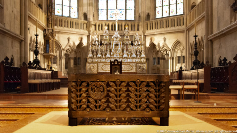 Carved altar table in the foreground of Regensburg Cathedral, aligned with the ornate high altar, choir stalls, and stained glass behind.