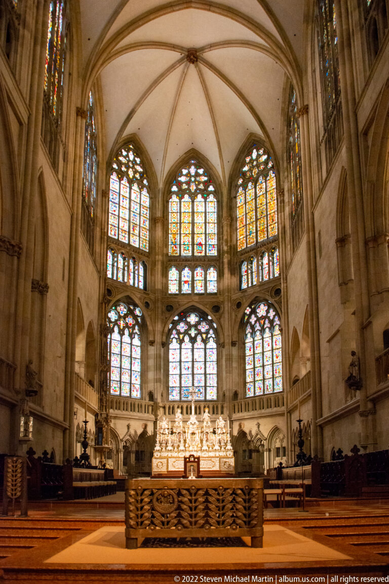 Choir of Regensburg Cathedral with stained-glass windows, high altar, stone vaulting, choir stalls, and carved altar rail.