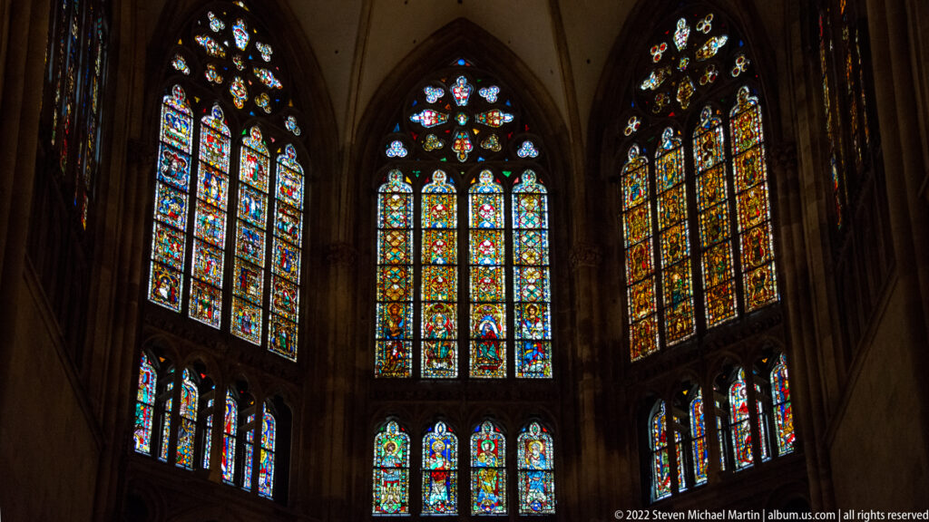 Multiple pointed stained-glass windows glowing in the darkened choir of Regensburg Cathedral.
