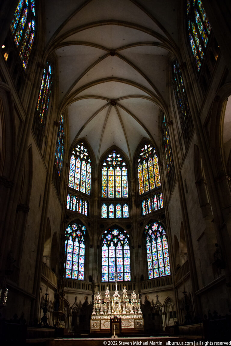 Upward view of Regensburg Cathedral choir with ribbed vaults, stained-glass windows, and illuminated high altar.