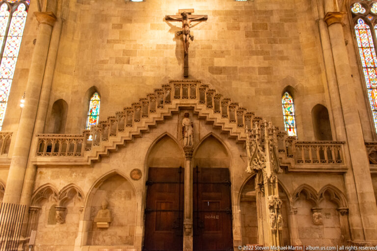 Two pointed wooden doors beneath an ornate Gothic stone monument, with sculpted figures, pinnacles, crucifix, and stained-glass windows.