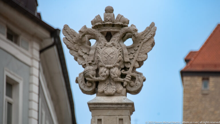 Carved stone double-headed eagle with spread wings, crown, chest shield, and ornate feathering on a pedestal.