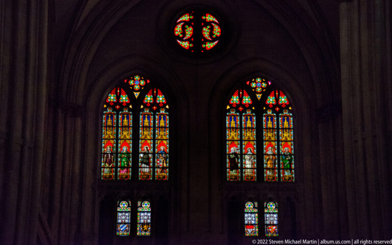 Two tall stained-glass Gothic windows beneath a circular colored-glass opening in a dark stone wall.