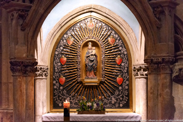 Crowned Virgin and Child statue centered in a radiating shrine with heraldic shields, flowers, and candles.