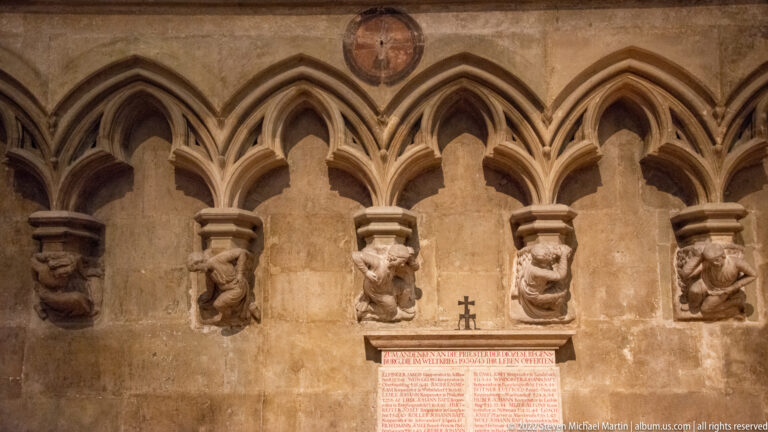 Five carved figural corbels beneath a row of pointed blind arches, with an inscription panel below.