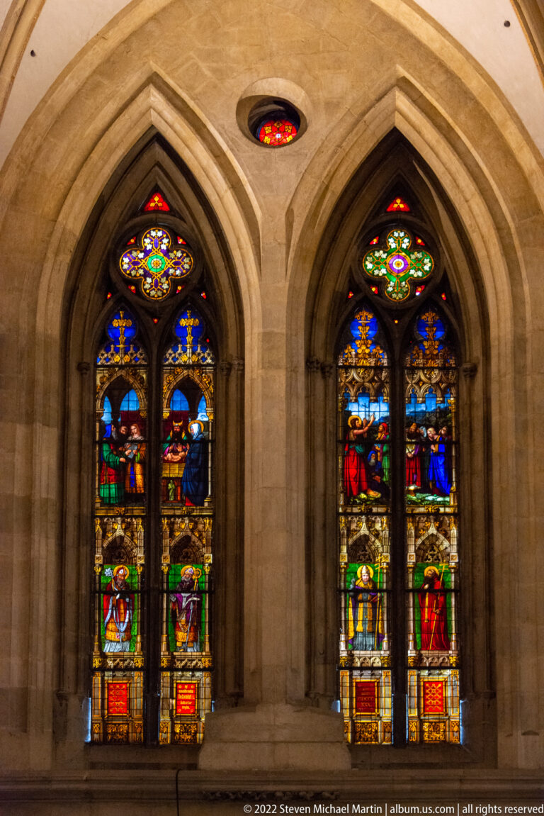 Two pointed stained-glass windows with stacked narrative scenes and small circular colored-glass openings above.