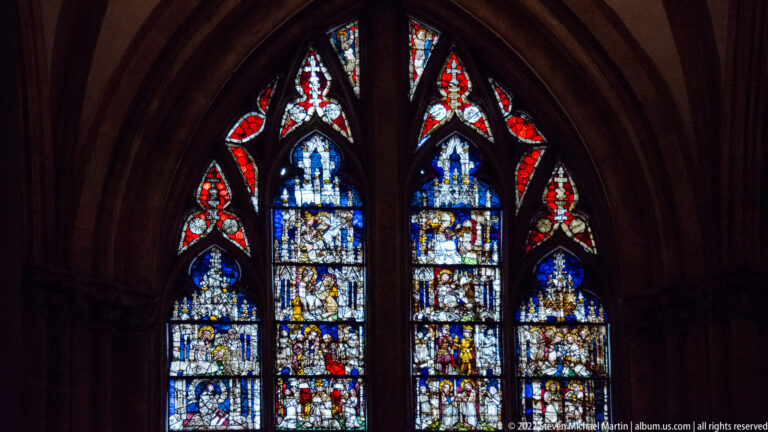 Two-lancet stained-glass window filled with small figurative scenes and red, white, and blue Gothic tracery.