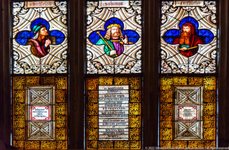 Three stained-glass portrait panels of bearded men above ornate inscription fields and decorative borders.