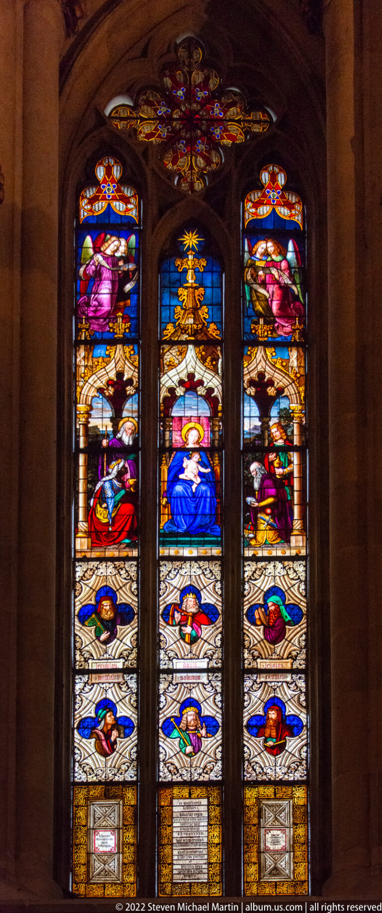 Three-panel stained-glass window with Virgin and Child at center, flanked by sacred figures and decorative scenes.