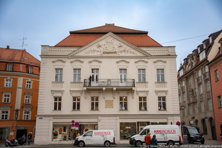 White historic building with triangular pediment, central balcony, symmetrical windows, and shops at street level.