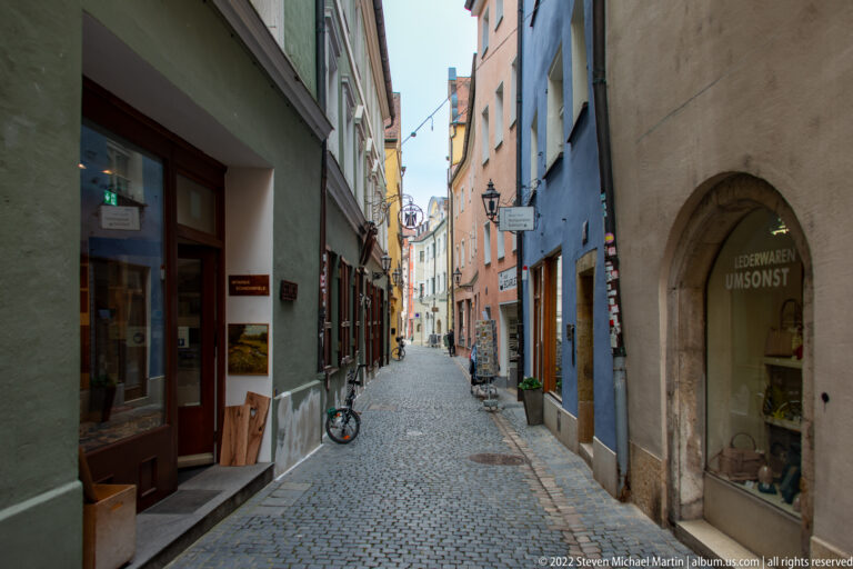 Narrow cobblestone lane lined with pastel historic buildings, small shops, signs, and bicycles in Regensburg.