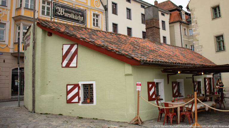 Low pale-green Wurstkuchl building with red-and-white shutters, tiled roof, outdoor tables, and surrounding old-town buildings.