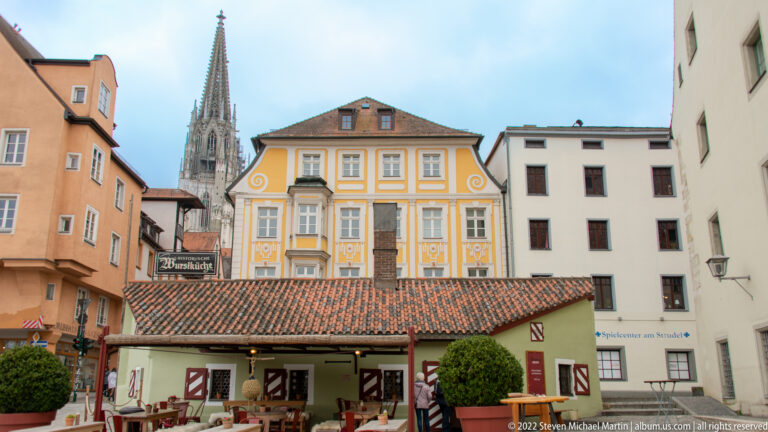 Wurstkuchl beneath a tiled roof with painted historic façades and a Regensburg Cathedral spire rising behind.