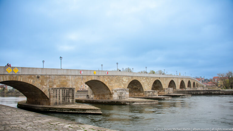 Long medieval stone bridge crossing the Danube with multiple arches, massive piers, cutwaters, and lamps along the parapet.