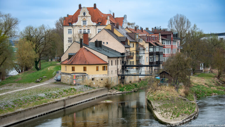 Historic buildings with tiled roofs and balconies lining a narrow river channel beside a grassy and tree-lined bank.