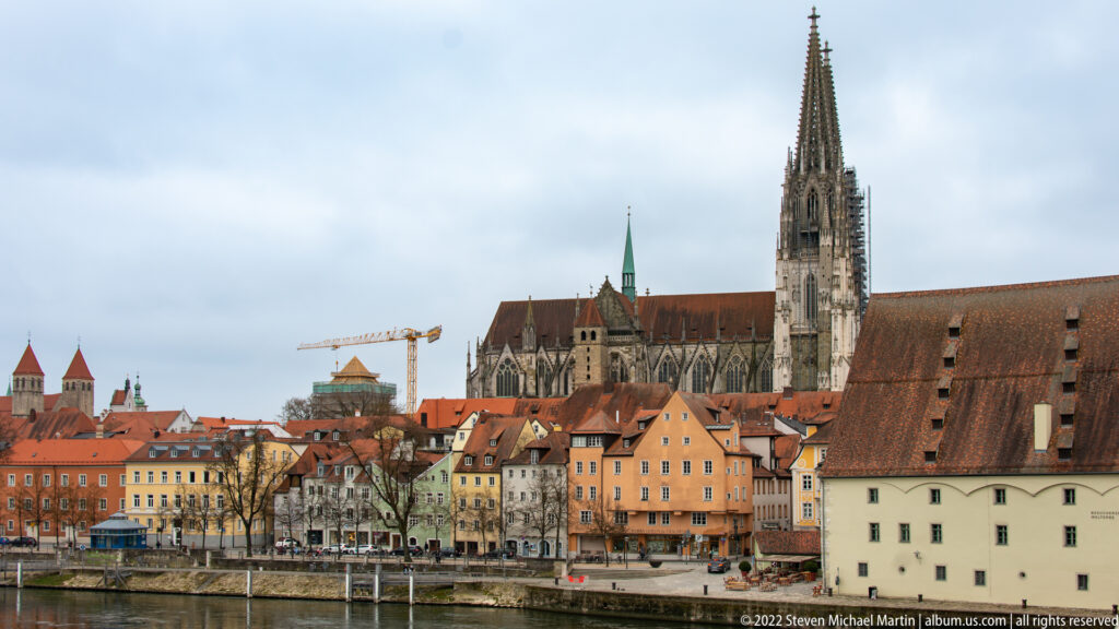 Regensburg old-town skyline along the Danube with the tall Gothic spire of St. Peter's Cathedral rising above red roofs.