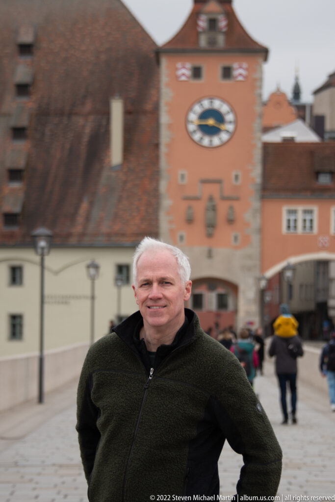 Man standing on Regensburg's Stone Bridge with the clock-faced Bridge Tower and pedestrians behind him.