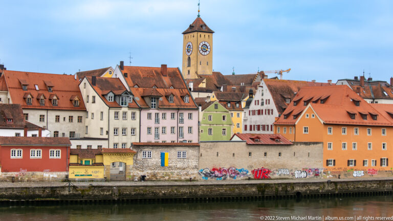 Colorful Regensburg riverfront with red roofs, varied gables, long riverside wall, and a tall square clock tower.