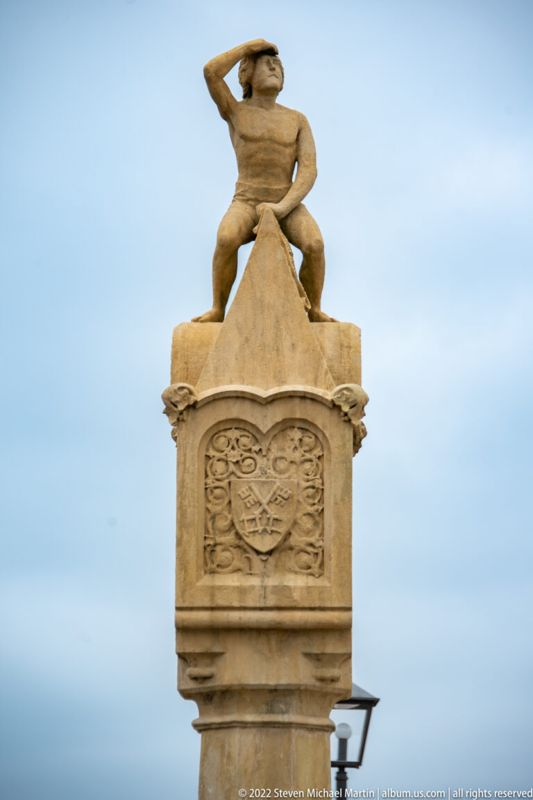 Small seated stone Bruckmandl figure shading its eyes atop a carved pedestal on Regensburg's Stone Bridge.