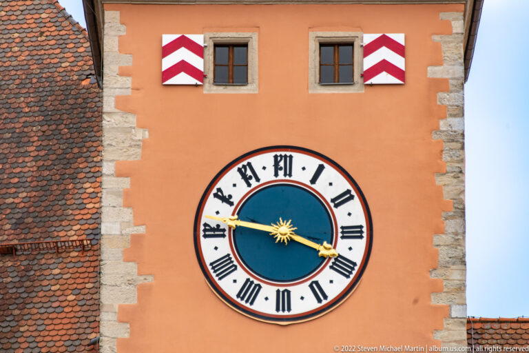 Large Roman-numeral clock with blue center and gilded hands on the orange façade of Regensburg's Bridge Tower.
