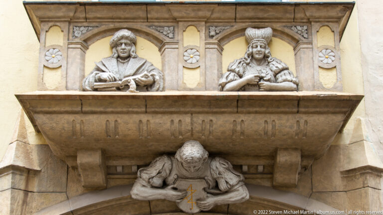 Stone architectural projection with two seated figures above and a bent figure beneath, framed by carved roundels and moldings.