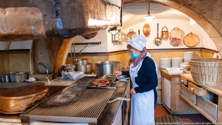 Cook tending sausages on a long open grill inside the compact Wurstkuchl kitchen, surrounded by copper pans and cookware.