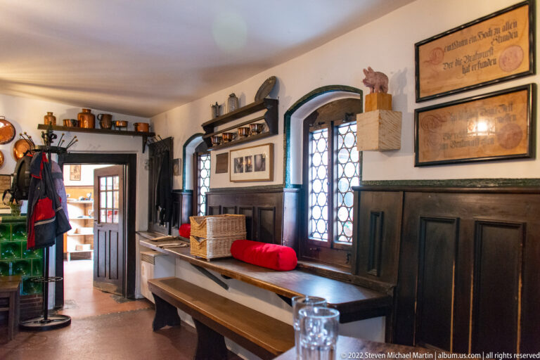 Traditional Regensburg tavern interior with dark wood paneling, communal table, leaded windows, framed prints, and copper cookware.