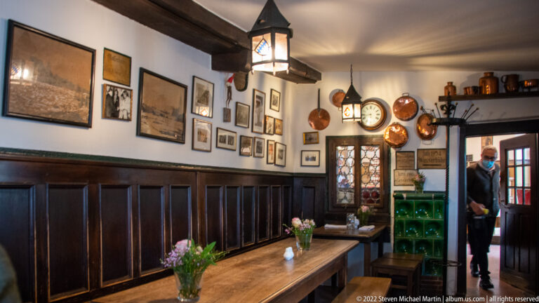 Long communal table in a wood-paneled tavern, with framed pictures, copper pans, clocks, bottles, and hanging lanterns.