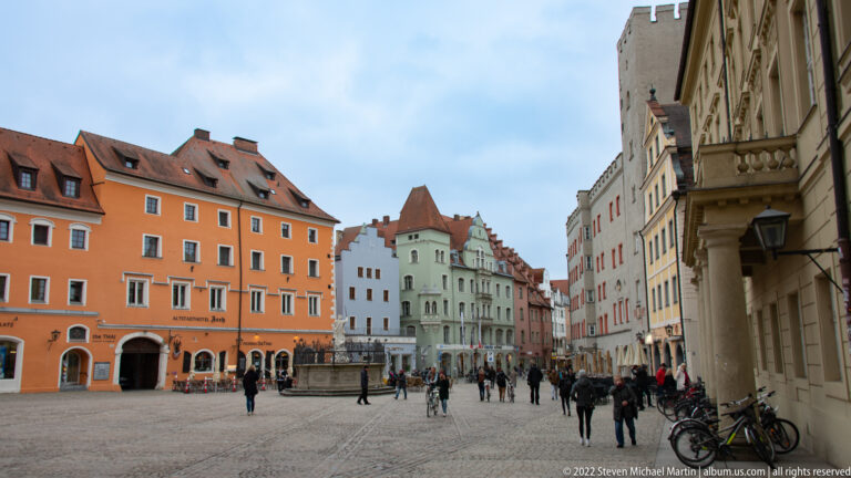 Cobbled Regensburg square with central stone fountain, pedestrians, bicycles, and colorful historic buildings with varied gables.