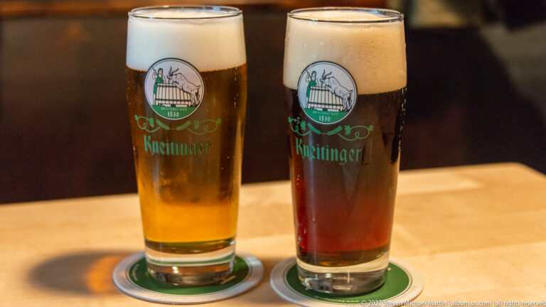 Two foamy Kneitinger beers, one golden and one dark amber, in branded glasses on coasters.