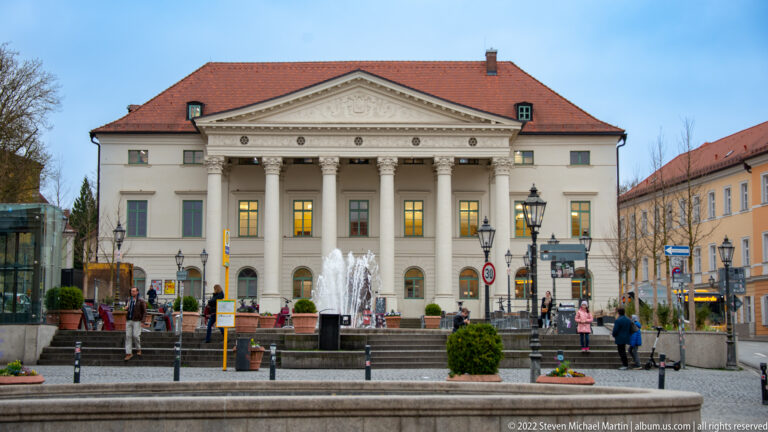 Neoclassical Theater Regensburg with columned portico and triangular pediment, fronted by steps, fountains, and pedestrians.