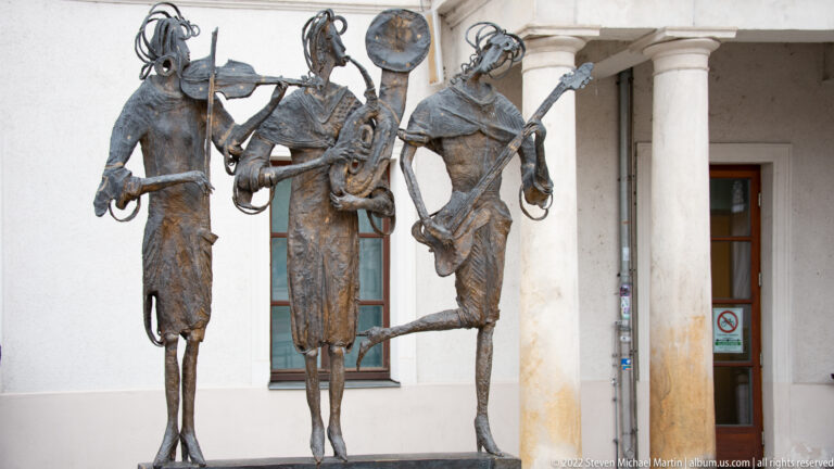 Bronze sculpture of three elongated costumed figures posing together outside a columned building in Regensburg.