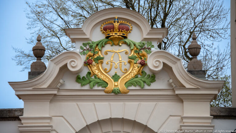Ornate gateway crest with gilded crowned monogram, green foliage, floral decoration, scrolls, and stone finials.
