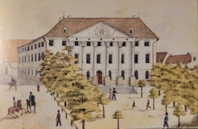 Historical illustration of a classical theater with columned portico and triangular pediment facing a landscaped public square.