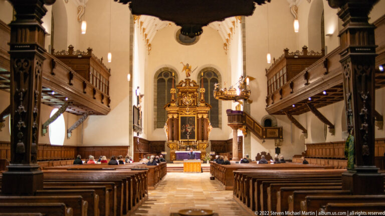 Neupfarrkirche nave with dark wooden side galleries, rows of pews, pale walls, high windows, and ornate altar.