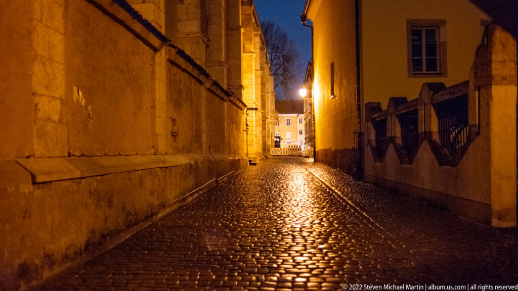 Narrow wet cobblestone lane at night, enclosed by high masonry walls and lit by warm streetlights.