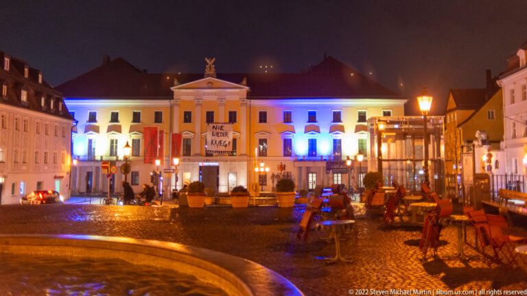 Illuminated Theater Regensburg at night across wet Bismarckplatz, with blue-white façade lighting, fountain, and outdoor seating.