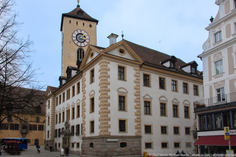 White historic civic building with rusticated corners and pediment, backed by a tall clock tower with circular clock face.