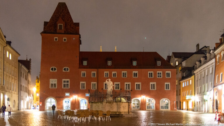 Red historic civic building with tall square tower and central stone fountain in a wet Regensburg square at night.
