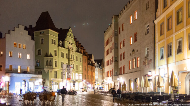 Regensburg old-town street at night with wet cobblestones, illuminated historic façades, gables, shopfronts, and outdoor tables.