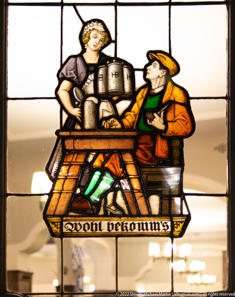 Stained-glass tavern scene with a woman carrying beer vessels toward a seated man in orange and green clothing.