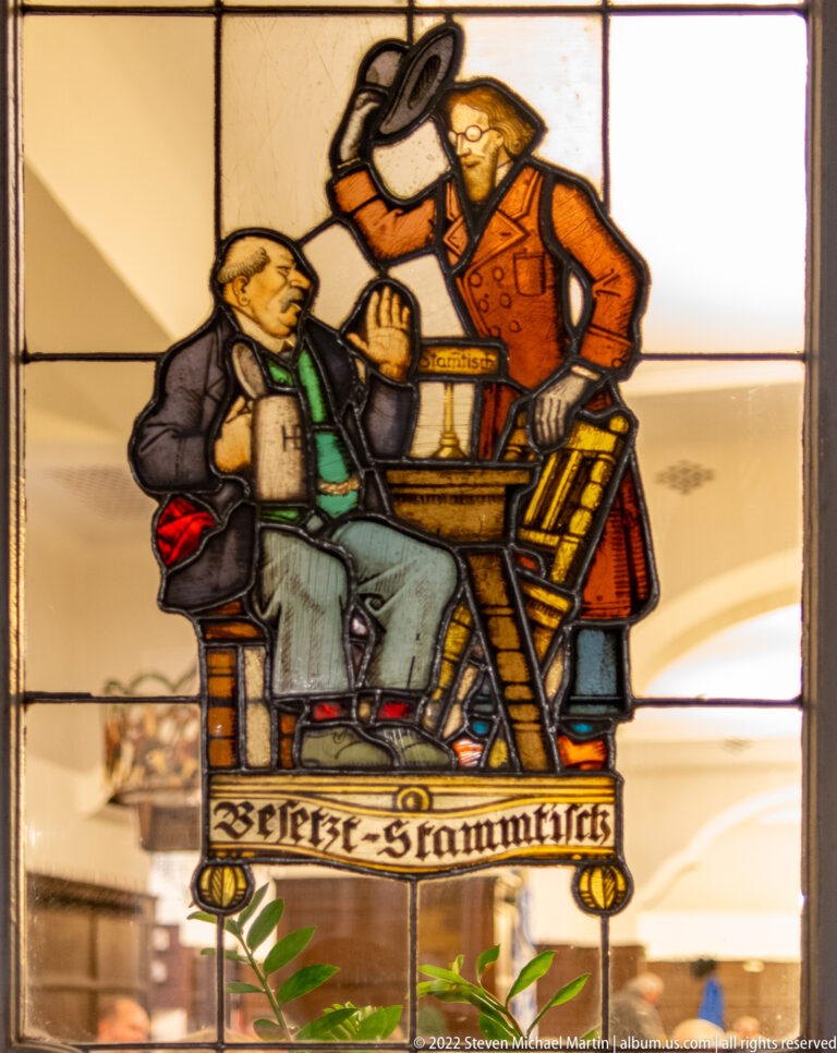 Stained-glass panel of two men at a small table, one seated with a mug and the other leaning over him.
