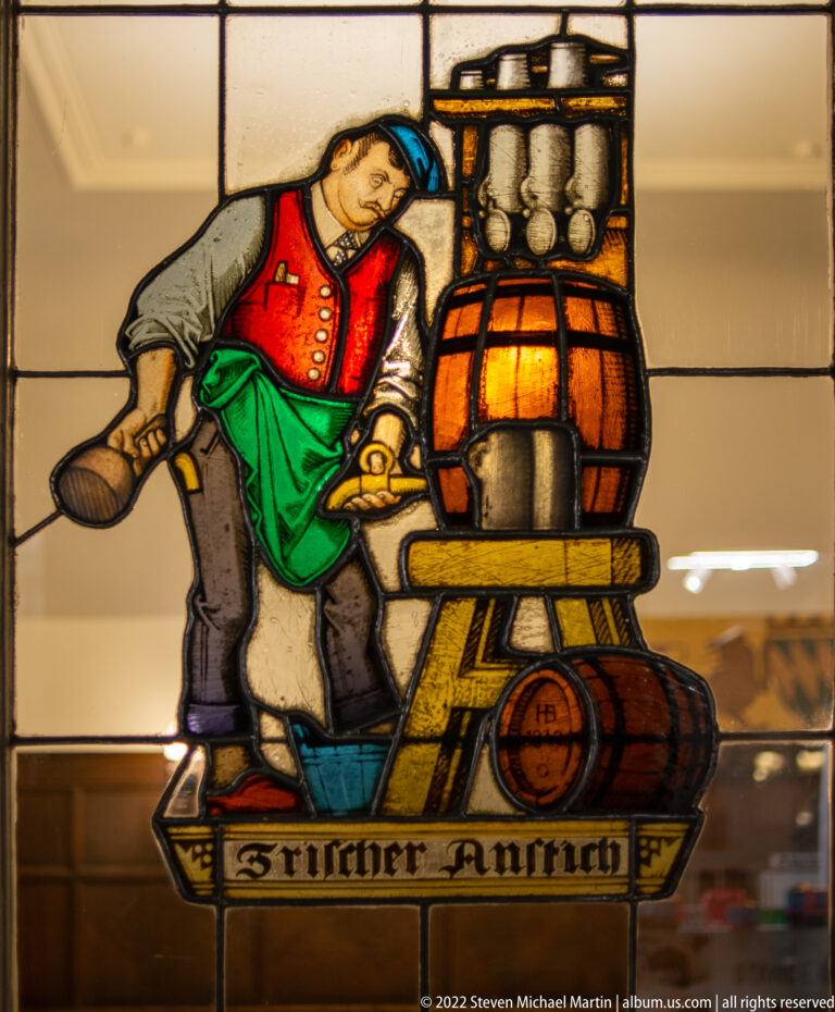 Stained-glass cellar worker leaning beside a large beer barrel with mugs stacked above and smaller casks below.