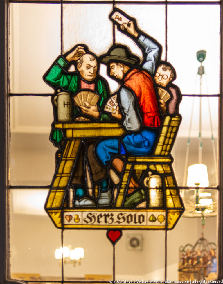 Stained-glass scene of several men crowded around a tavern table with a large drinking vessel.