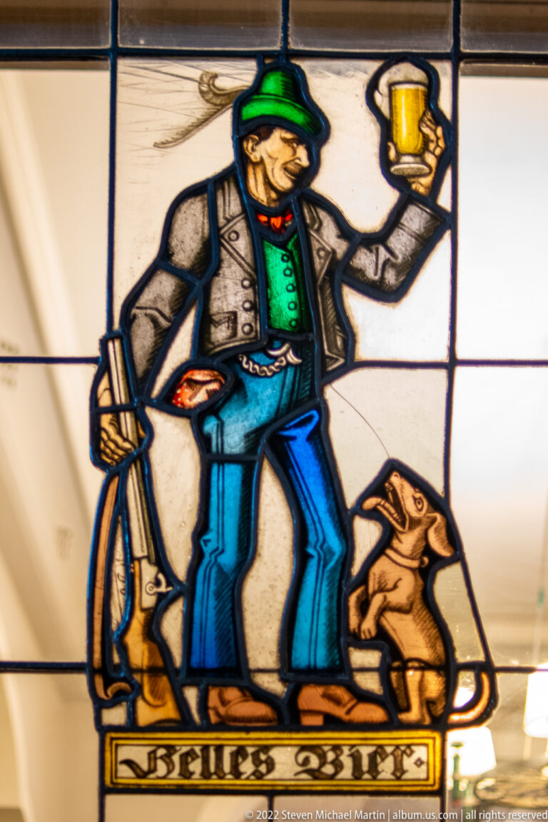 Stained-glass figure in green cap raising a beer mug beside a seated dog.